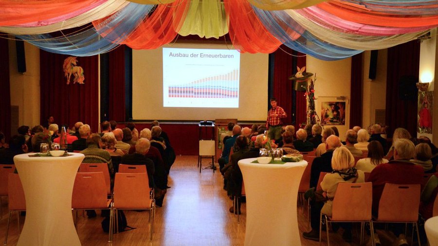 Zu sehen ist der große Saal der Friesenhalle mit Publikum und einer Leinwand, auf der eine Folie mit dem Titel »Ausbau der Erneuerbaren« gezeigt wird.