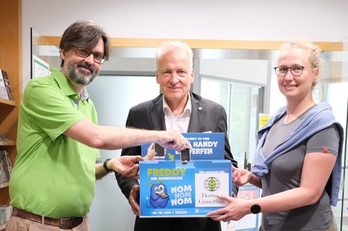 Ein altes Handy für die Sammelbox Jan Bertram (Referent für Digitales und IT), Peter Münster (1. Bürgermeister) und Rike Schiele (3. Bürgermeisterin) sind mit einer Handy-Sammelbox zu sehen, die im Rathaus aufgestellt wird. Jan Bertram wirft gerade ein altes Nokia-Handy in die Sammelbox.
